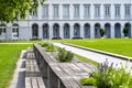 Benches and tables with flowers arranged in a row in the park, in the background a large building with arched windows.