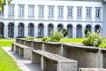 Benches and tables with flowers arranged in a row in the park, in the background a large building with arched windows.