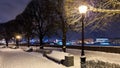 Benches under snow along alley with trees and street lights on embankment. Winter cityscape at twilight. Saint Petersburg, Russia.