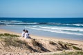 An adult couple looking over the ocean while sitting on sand dunes on a sunny day. Bendalong Beach