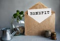 BENEFIT text is written on white paper on an antique envelope, which lies on the table along with a stack of coins, a glass jar of Royalty Free Stock Photo