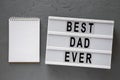 `Best dad ever` words on lightbox, blank notepad over gray background, top view. Overhead, from above, flat lay