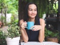 Beutiful Asian woman with long hair sitting in the garden, holding blue cup of coffee, smiling and looking  away.. relaxing with Royalty Free Stock Photo