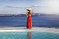 A beutiful woman in red dress and with hat stands on the edge of a swimming pool