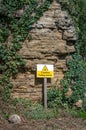 Beware of falling rock warning sign with triangle picture and alert message in yellow and black. Stood in front of cliff face.