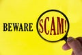 Beware scam text on yellow cover with hand holding magnifying glass. Scamming and fraud concept