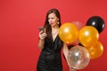 Bewildered young woman in little black dress holding air balloons using mobile phone while celebrating isolated on red Royalty Free Stock Photo