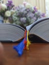 Bible book with yellow, red and blue ribbons marker on the wooden table with blur flowers bouquet background. Christianity.