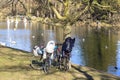 Bicycles in the park by the pond in which birds float. People on the other side launch model sailboats