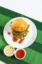 Big beef steak burger with vegetables and herbs on white plate on green bamboo placemat Royalty Free Stock Photo