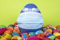 Big blue egg with face mask between painted easter eggs before gren background Royalty Free Stock Photo