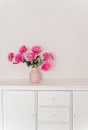 Big bouquet of fuchsia peonies in a pink vase in a bright room are on a white chest of drawers. cozy house