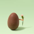 Big chocolate egg with female hand holding small flower pink carnation minimal arrangement. Vivid Green background