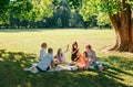 Big family sitting on picnic blanket in city park linden treeb during weekend Sunday sunny day. They are chatting and eating Royalty Free Stock Photo