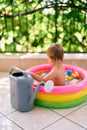 Big garden watering can stands against the background of a toddler sitting in an inflatable mini-pool