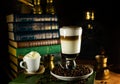 Big glass with latte stand in coffee beans. Atmosphere of vinatge library with old books around and grinder on dark background. Royalty Free Stock Photo