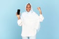 Big luck. Excited black muslim woman holding blank smartphone and shaking fist, exclaiming with excitement