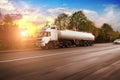 Big metal fuel tanker truck shipping fuel on the countryside road in motion with trees against sky with sunset