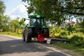 Big modern industrial tractor machine cutting green grass with mowing equipment along country roadside. Road lawn mower