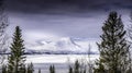 Big mountain with clouds at the top, behind big frozen lake with pine trees in foreground Royalty Free Stock Photo
