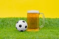 Big mug filled with cold beer with foam, and miniature soccer ball stand on grass of artificial lawn on yellow Royalty Free Stock Photo