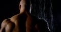 Big muscular jock is standing in shower under water stream, back view in darkness