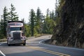 Big rig American bonnet semi truck with covered cargo moving around huge cliff on the winding road Royalty Free Stock Photo