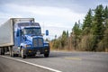 Big rig blue classic powerful semi truck transporting frozen cargo in refrigerated semi trailer running on the road with trees on