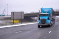 Big rig blue classic semi truck with covered dry van semi trailer and high exhaust pipes is moving along multi-lane highway in sn Royalty Free Stock Photo