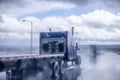 Big rig blue classic semi truck with flat bed semi trailer moving on the wet raining road behind another semi truck with rain dust