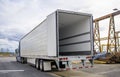 Big rig blue semi truck with open door semi trailer standing at warehouse parking lot at industrial area waiting for the load for