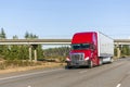 Big rig bright red semi truck with dry van semi trailer running on wide highway with concrete bridge over the road Royalty Free Stock Photo