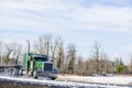 Big rig green American bonnet semi truck with flat bed semi trailer driving on the winter snowy road with wet dust surface Royalty Free Stock Photo