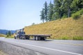 Big rig semi truck with crane application on the frame transporting empty flat bed semi trailer running on the winding road with Royalty Free Stock Photo