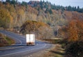 Big rig semi truck transporting goods in dry van semi trailer running on the winding road with autumn trees on the hills