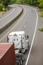 Big rig white low cab semi truck transporting container on turning road with green trees on the sides