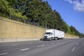 Big rig white semi truck with dry van semi trailer running on the wide multiline highway road with concrete wall and trees on the Royalty Free Stock Photo