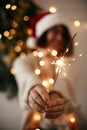 Big sparkler burning in hand of stylish girl in santa hat on background of modern christmas tree light in dark room. Woman with Royalty Free Stock Photo