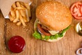 Big tasty burger on the wooden table with becon and french fries. Delicious dinner with cheese, tomatoos and salad. Fast food with
