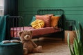 Big teddy bear and an open book on a green, velvet pouf in front of a metal frame bed in child`s bedroom interior. Real