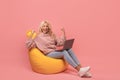 Big win and success concept. Excited lady sitting on beanbag chair with laptop and raising hands up, pink background