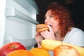 Big woman eat fast food. Red hair fat girl looking inside refrigerator with burger. Unhealthy and healthy food concept with plus Royalty Free Stock Photo