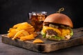 Big yummy burger with cheese, fries and a glass of cola on a wooden board on a black background, angle view. Royalty Free Stock Photo