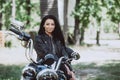 Biker girl in a leather jacket on a motorcycle looks at the camera on a summer sunny day on a green background. Soft focus