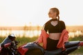 A biker girl next to a motorcycle by a pond in the orange rays of the setting sun, a selective focus. Copy space