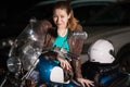 Biker girl portrait, young heavily-built woman standing near her motorcycle and white helmet in dark parking