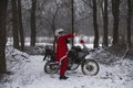 A biker in a helmet puts on a Santa Claus costume.. Winter forest with falling snow. Touring motorcycle in the background. The Royalty Free Stock Photo
