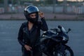 Biker woman in black leather jacket and full face helmet stands near stylish sports motorcycle. Urban parking, sunset in Royalty Free Stock Photo