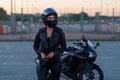 Biker woman in black leather jacket and full face helmet stands near stylish sports motorcycle. Urban parking, sunset in Royalty Free Stock Photo