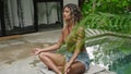Multiracial woman meditates in tranquil poolside setting. Bikini-clad, sits cross-legged, eyes closed, amidst plants